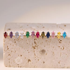 May include: A collection of colorful gemstone earrings displayed on a textured, light-colored surface. Each earring features a marquise-cut gemstone in a different color, including red, purple, blue, and green. The earrings are set in gold.