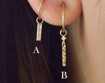 May include: Two gold hoop earrings with dangling charms. The left earring has a charm with a row of clear cubic zirconia stones. The right earring has a charm with a single clear cubic zirconia stone and a textured gold bar.