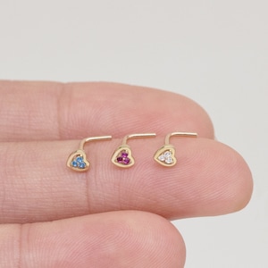 May include: Three gold heart-shaped nose rings with different colored gemstones. The rings are on a finger, and the gemstones are blue, pink, and white.