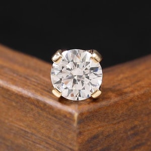 May include: A round brilliant cut clear gemstone set in a four-prong gold metal stud earring. The stone features multiple facets that reflect light, and the setting is secured with four small metal prongs holding the gem in place securely.