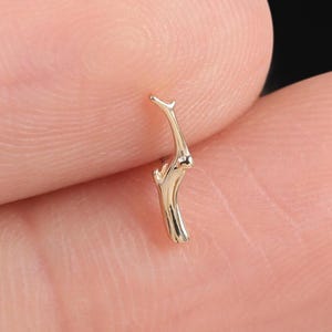 May include: A small, gold-colored twig-shaped earring. The earring has a detailed, textured surface and a curved design. The earring is shown against a blurred background.
