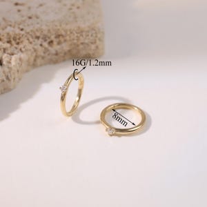 May include: Two gold-colored hoop earrings with a small, clear gemstone. One earring is open, and the other is closed. The open earring is labeled "16G/1.2mm". The closed earring is labeled "8mm".