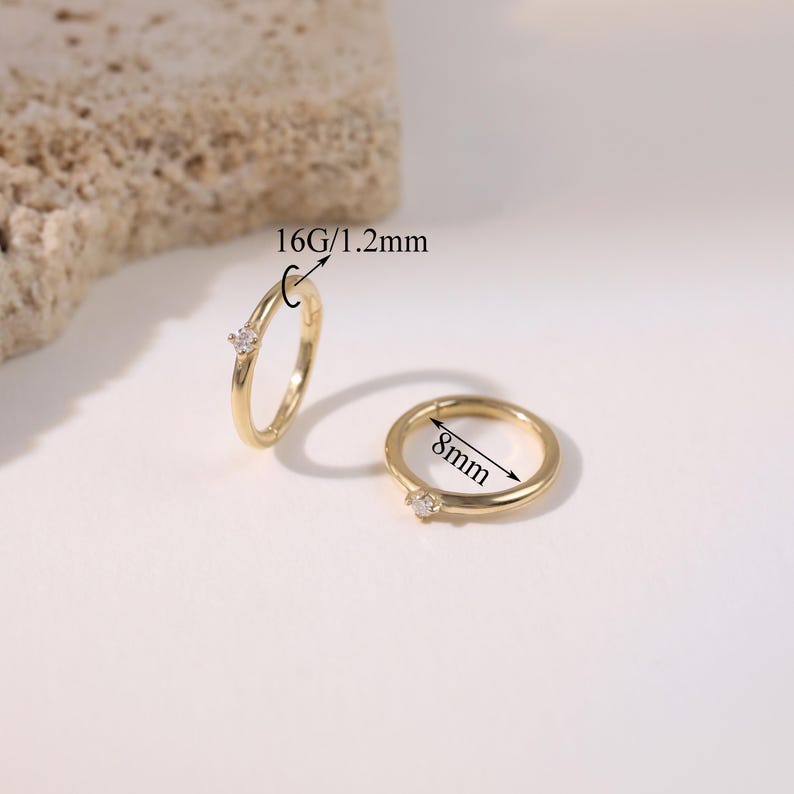 May include: Two gold-colored hoop earrings with a small, clear gemstone. One earring is open, and the other is closed. The open earring is labeled "16G/1.2mm". The closed earring is labeled "8mm".