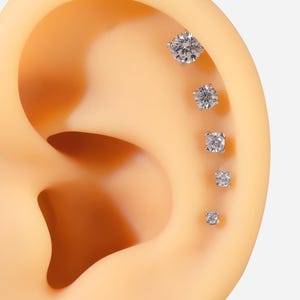 Pu&ograve; includere: Cinque orecchini a perno d'argento con pietre di zircone cubico trasparenti di diverse dimensioni. Gli orecchini sono disposti in linea sull'orecchio di un manichino.