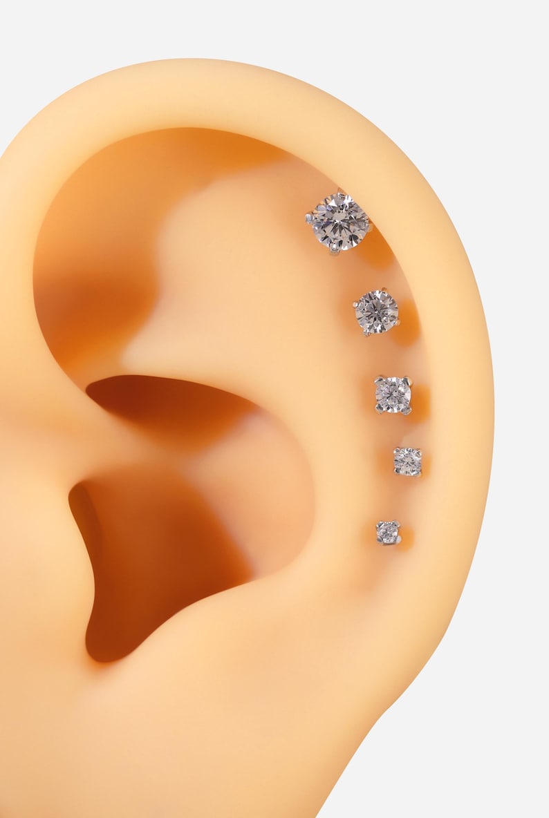 Pu&ograve; includere: Cinque orecchini a perno d'argento con pietre di zircone cubico trasparenti di diverse dimensioni. Gli orecchini sono disposti in linea sull'orecchio di un manichino.