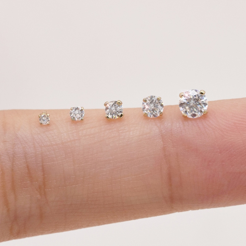 May include: Five different sizes of clear round cubic zirconia stud earrings in gold settings. The earrings are lined up in order of size from smallest to largest.