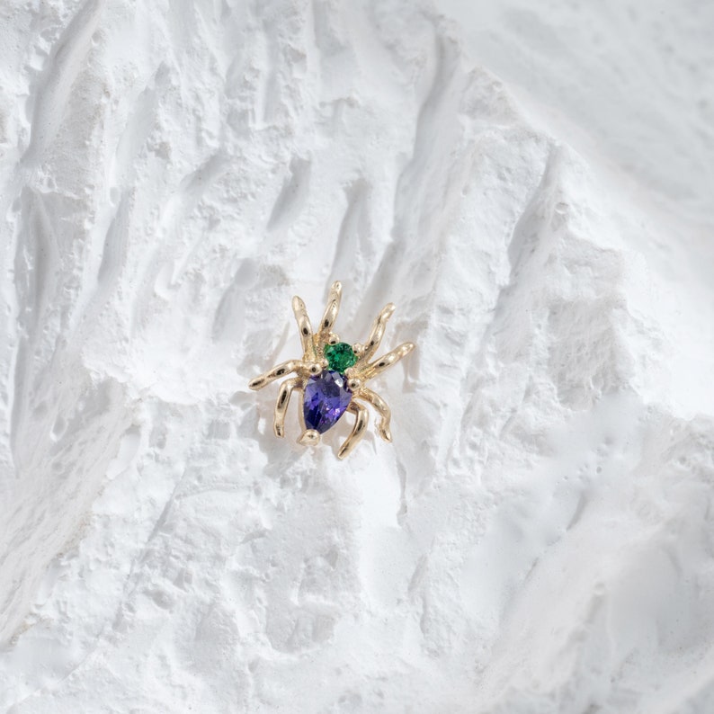 May include: A gold spider brooch with a purple and green gemstone accent.