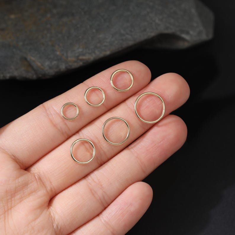 May include: A collection of six gold-toned circular rings of varying sizes arranged in a cluster. Each ring features a smooth, thin, continuous metal band with a simple, circular design, showcasing a polished finish that reflects light.