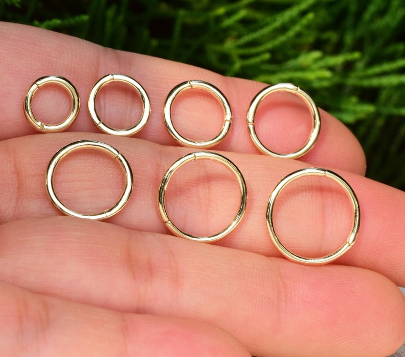 May include: A collection of seven circular gold-toned metal rings arranged in two rows. Each ring features a smooth, polished surface and a single visible seam where the ends meet, creating a simple, continuous hoop design in varying sizes.