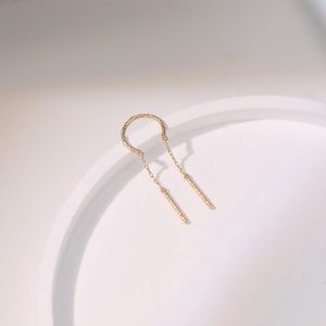 May include: A gold-colored earring with a textured, curved top and two dangling, textured bars. The earring is set against a white background, with soft shadows. The design is minimalist and elegant.