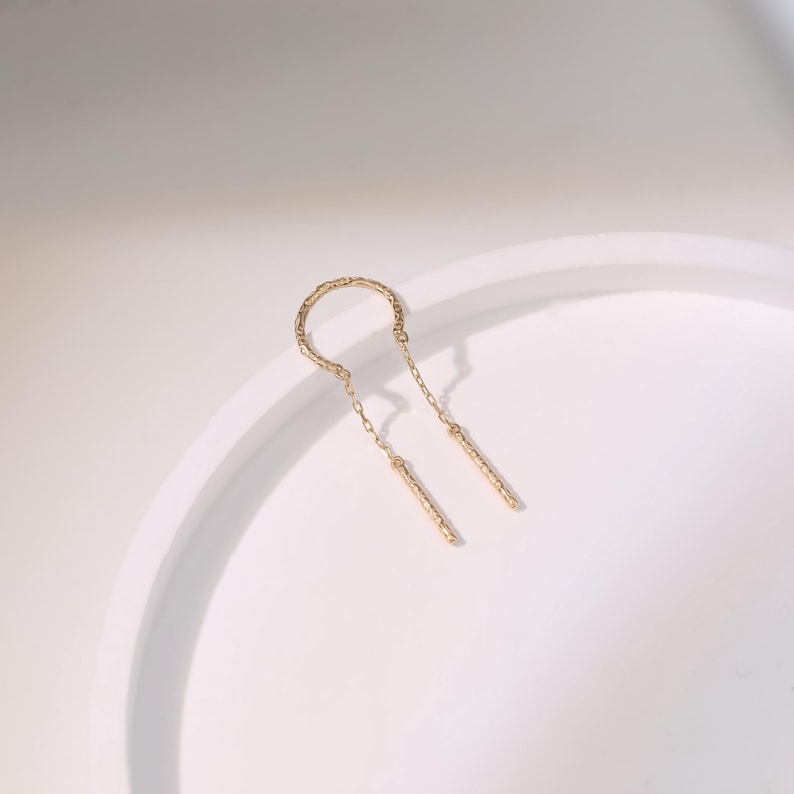 May include: A gold-colored earring with a textured, curved top and two dangling, textured bars. The earring is set against a white background, with soft shadows. The design is minimalist and elegant.