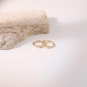 May include: Two delicate gold rings, each featuring a small, clear gemstone. The rings are displayed on a white surface, with a textured, light-brown stone in the background. The rings are simple and elegant.