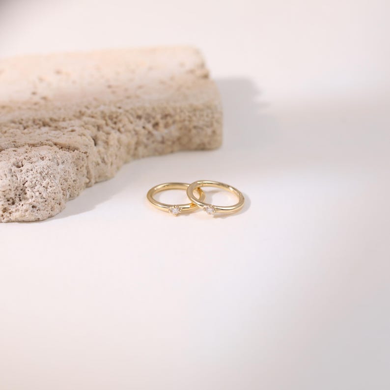 May include: Two delicate gold rings, each featuring a small, clear gemstone. The rings are displayed on a white surface, with a textured, light-brown stone in the background. The rings are simple and elegant.