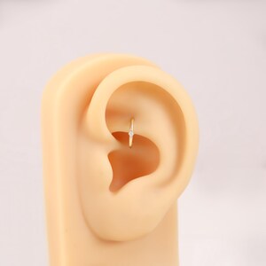 May include: A small, gold-colored hoop earring with a single, clear gemstone. The earring is set in the upper ear cartilage of a mannequin. The earring is a simple, elegant design.