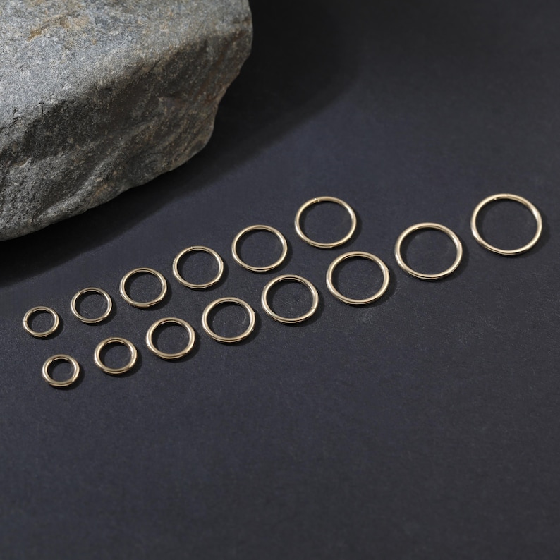 May include: A collection of fifteen gold-toned metal jump rings arranged in a scattered pattern. The rings vary in size, featuring a thin, circular wire construction with a smooth, polished finish, presented as individual open-ended connectors.