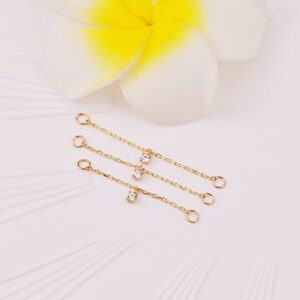 May include: Three gold chains with small clear gemstones hanging from them. The chains have jump rings at both ends.