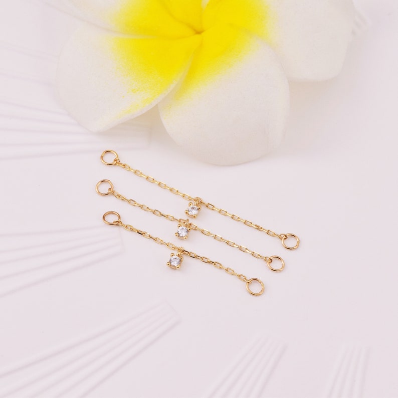 May include: Three gold chains with small clear gemstones hanging from them. The chains have jump rings at both ends.