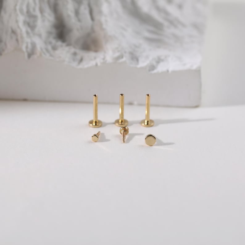 K&ouml;nnte beinhalten: Eine Sammlung goldfarbener K&ouml;rperschmuckst&uuml;cke, darunter drei flachr&uuml;ckenbesetzte Ohrstecker, drei Stifte und ein kleines, dekoratives St&uuml;ck. Der Schmuck ist auf einer wei&szlig;en Oberfl&auml;che angeordnet, mit einem weichen, strukturierten Hintergrund.