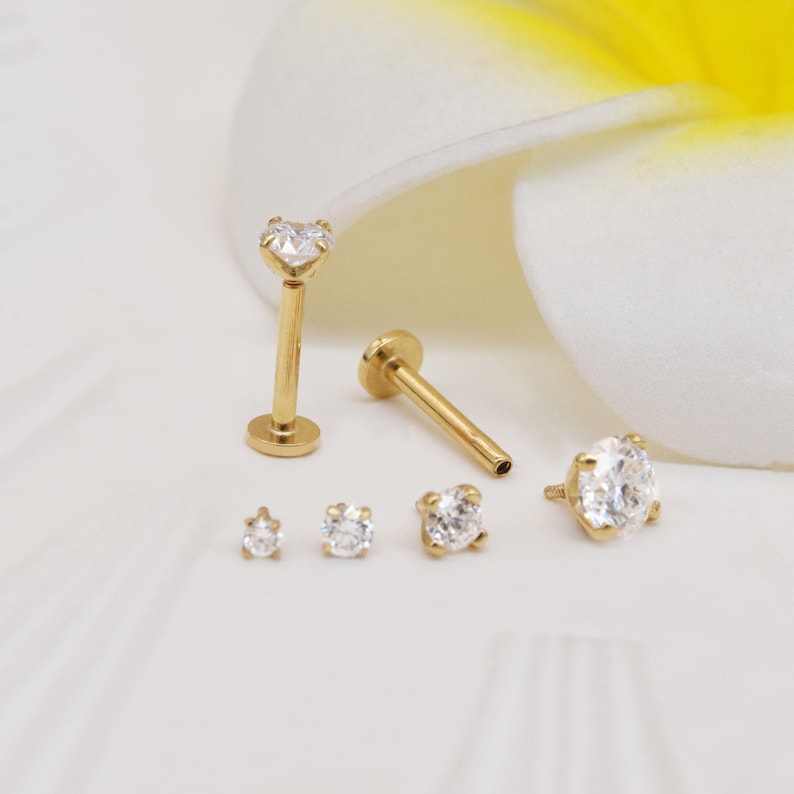 May include: A set of five gold-toned body jewelry pieces with clear cubic zirconia stones. The set includes two labret studs, one with a heart-shaped stone and one with a round stone, and three smaller round stones.