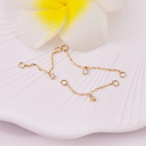 May include: Three gold chains with small clear gemstones hanging from each end. The chains are on a white surface.