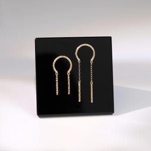 May include: A pair of gold-colored earrings displayed on a black surface. The earrings feature a textured, open-circle design at the top, with a dangling bar element. One earring has a chain, while the other has a solid bar.