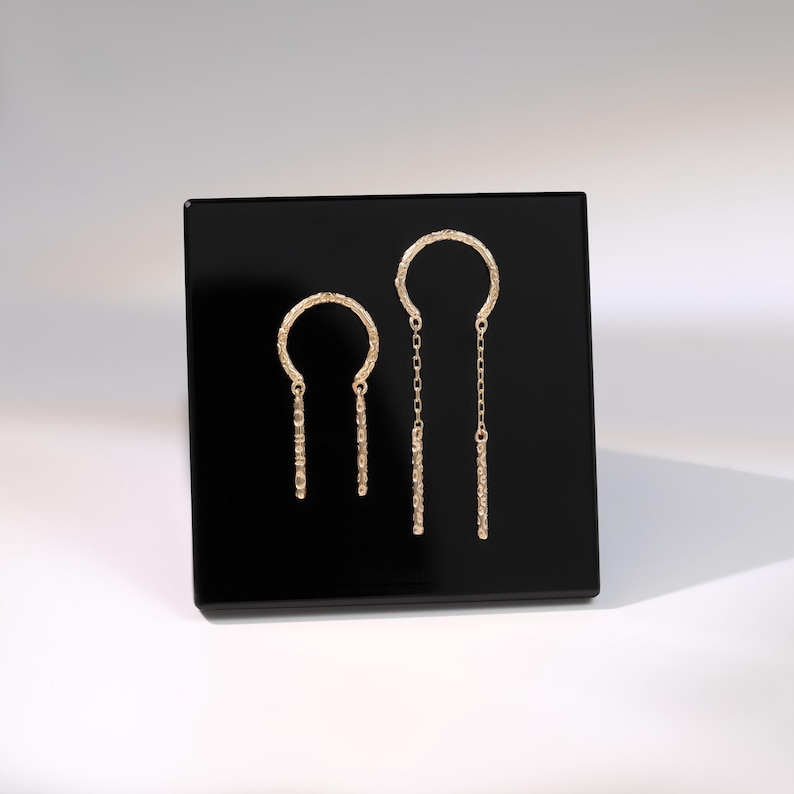 May include: A pair of gold-colored earrings displayed on a black surface. The earrings feature a textured, open-circle design at the top, with a dangling bar element. One earring has a chain, while the other has a solid bar.