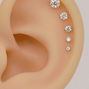 May include: Close-up of a person's ear with five small, clear, round gemstones set in gold studs. The gemstones are arranged in a line, starting at the top of the ear and going down.