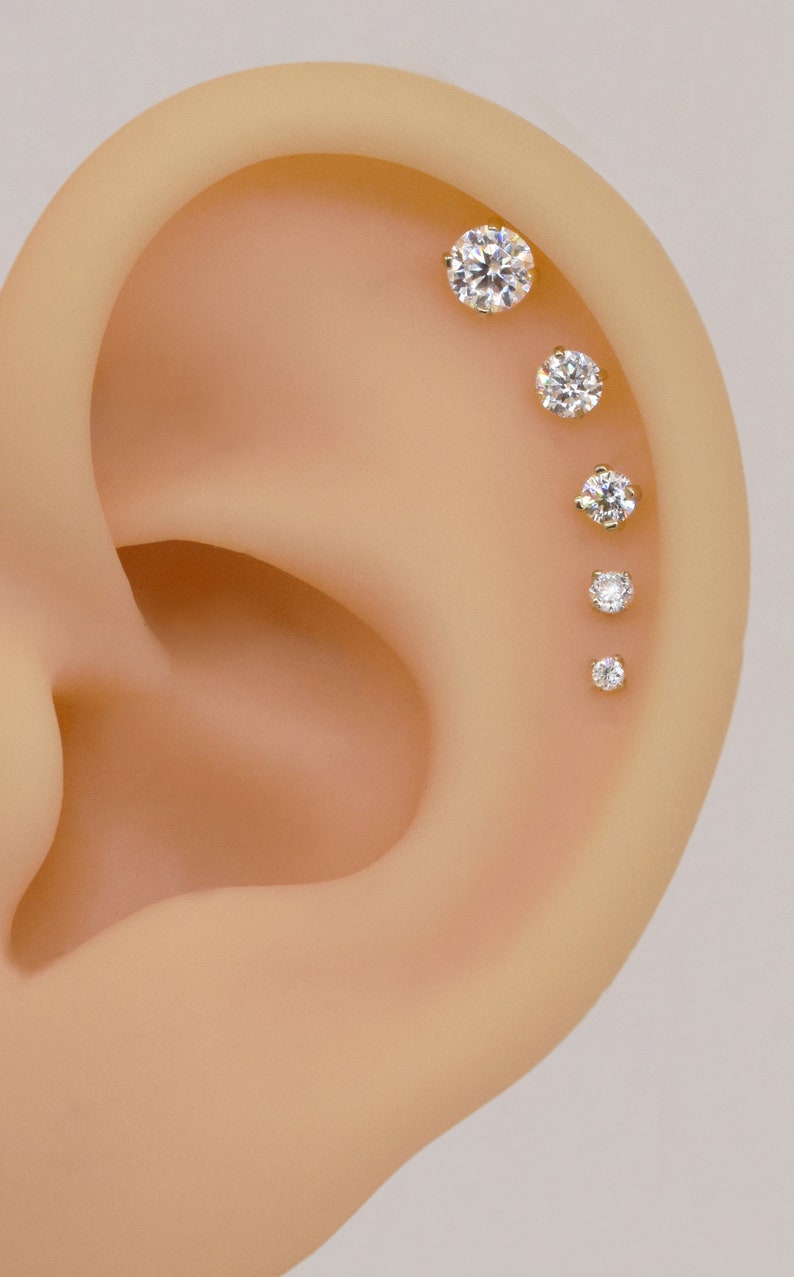 May include: Close-up of a person's ear with five small, clear, round gemstones set in gold studs. The gemstones are arranged in a line, starting at the top of the ear and going down.