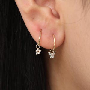 May include: Gold hoop earrings, one with a flower charm and the other with a butterfly charm, both adorned with small, clear gemstones. The earrings are displayed on an ear, showcasing their delicate design and sparkle.