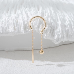 以下が含まれることがあります： ゴールドカラーのイヤーカフ。チェーン、垂直のバー、ファセット加工された球体がぶら下がっています。シンプルでエレガントなデザインのジュエリーで、白いテクスチャの背景に映えます。