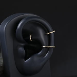 May include: Three thin, polished gold-tone hoop earrings displayed on a dark, matte ear-shaped model. One small hoop is positioned in the upper cartilage, another in the conch area, and a third hoop is placed in the lower cartilage section.