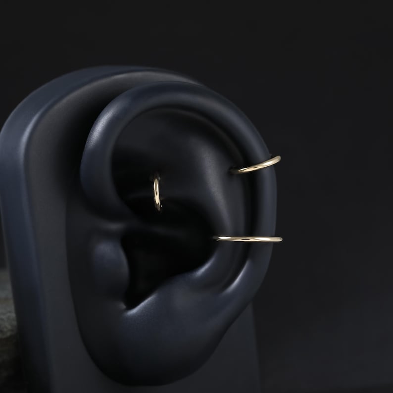 May include: Three thin, polished gold-tone hoop earrings displayed on a dark, matte ear-shaped model. One small hoop is positioned in the upper cartilage, another in the conch area, and a third hoop is placed in the lower cartilage section.