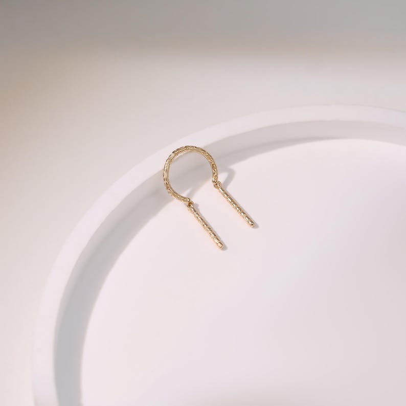 May include: A gold-colored earring with a textured, U-shaped top and two dangling, textured bars. The earring is set against a white background, highlighting its delicate design and craftsmanship. This piece of jewelry is suitable for everyday wear or special occasions.