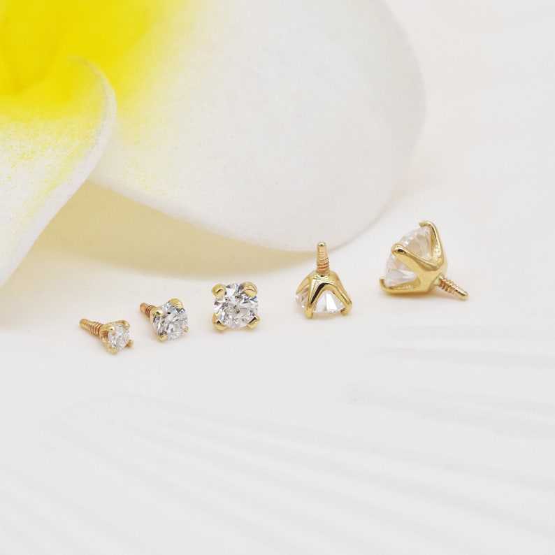May include: Four gold body jewelry pieces with clear gemstones. The pieces are arranged in a row from smallest to largest. The largest piece has a prong setting.