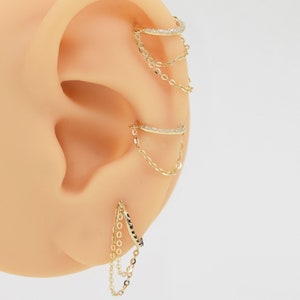 May include: Gold ear cuff with a chain dangling from it. The ear cuff is set with small clear stones. The chain is made of small gold links.