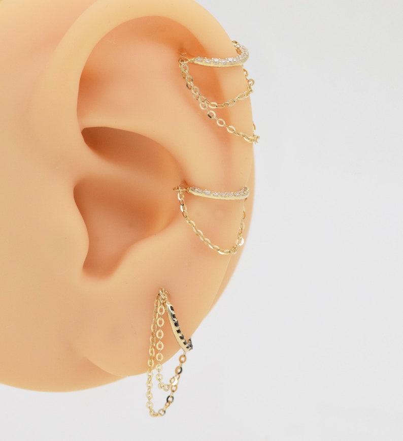 May include: Gold ear cuff with a chain dangling from it. The ear cuff is set with small clear stones. The chain is made of small gold links.