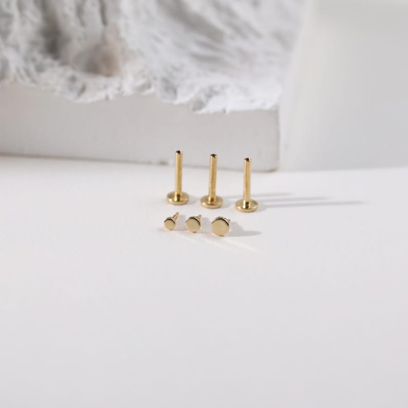 K&ouml;nnte beinhalten: Sechs goldfarbene Lippen- oder Ohrstecker sind auf einer wei&szlig;en Oberfl&auml;che angeordnet. Drei Stecker haben einen Stab und eine flache, runde Oberseite. Die anderen drei Stecker sind flach und rund. Die Stecker sind in verschiedenen Gr&ouml;&szlig;en.