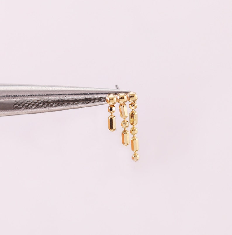 May include: Gold chain earring with a dangling design featuring a series of round and rectangular beads.