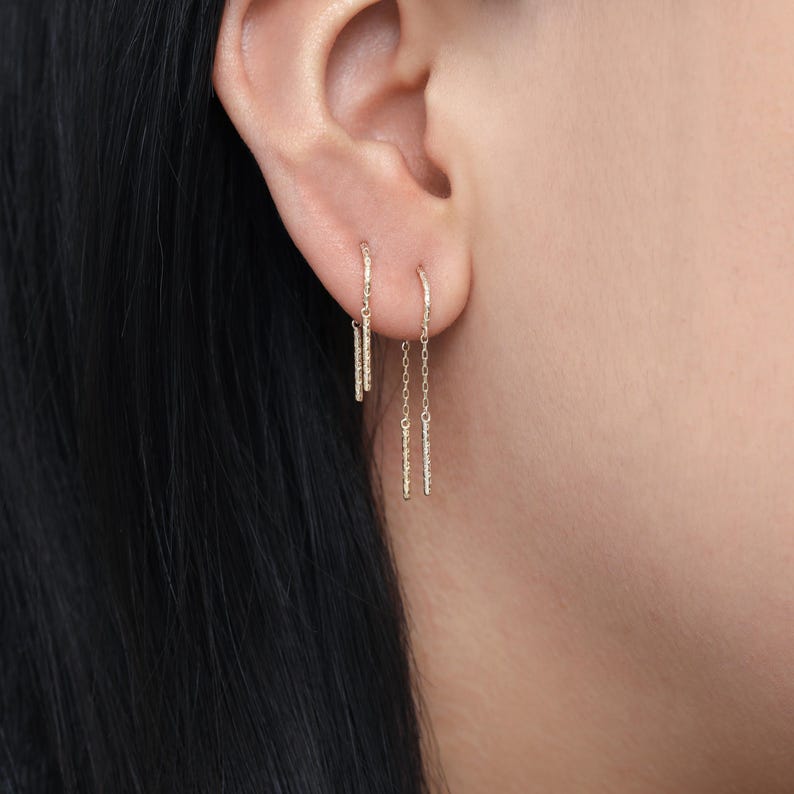 May include: A close-up of two gold earrings. One earring features a small hoop with a dangling bar, while the other has a chain with a bar. The earrings are worn on a person's ear, with the focus on the jewelry.