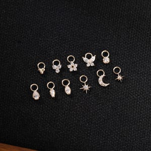 May include: A collection of small, gold-colored charms with various designs, including flowers, stars, and teardrops. Each charm features a small loop for attachment. The charms are set against a dark, textured background.