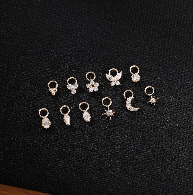 May include: A collection of small, gold-colored charms with various designs, including flowers, stars, and teardrops. Each charm features a small loop for attachment. The charms are set against a dark, textured background.