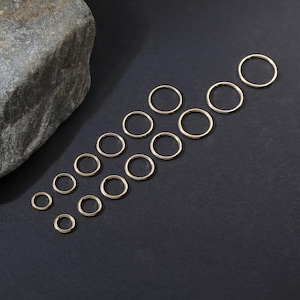 May include: A collection of fifteen gold-toned metal jump rings arranged in a staggered pattern. The rings vary in diameter, featuring a smooth, circular wire construction. These jewelry components are displayed on a dark, flat surface.
