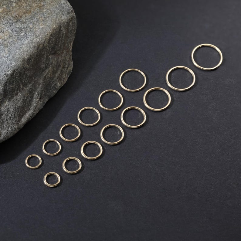 May include: A collection of fifteen gold-toned metal jump rings arranged in a staggered pattern. The rings vary in diameter, featuring a smooth, circular wire construction. These jewelry components are displayed on a dark, flat surface.
