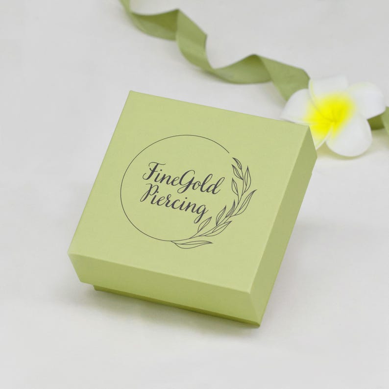 May include: A square, light green box with the text "FineGold Piercing" in a circular design. A green ribbon and a white flower with a yellow center are in the background. The box is likely for jewelry.