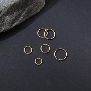 May include: A collection of six gold-toned metal jump rings of varying diameters arranged on a dark surface. The rings are circular, thin, and feature a smooth, polished finish. These jewelry components are displayed in different sizes.