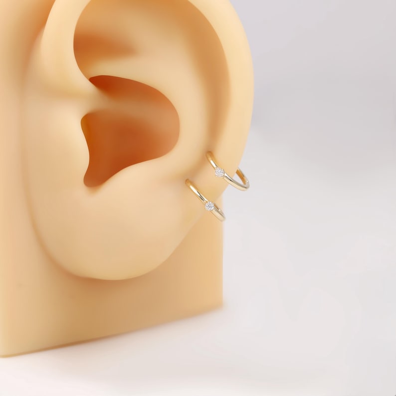 May include: Two gold-colored hoop earrings, each with a small, clear gemstone, are displayed on a tan ear model. The earrings are simple and elegant, with a minimalist design. The background is a plain white.