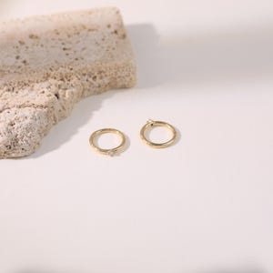 May include: Two gold hoop earrings with small, clear gemstones. One earring has a gemstone at the clasp, and the other has a gemstone in the center. The earrings are on a white surface with a textured, beige stone in the background.