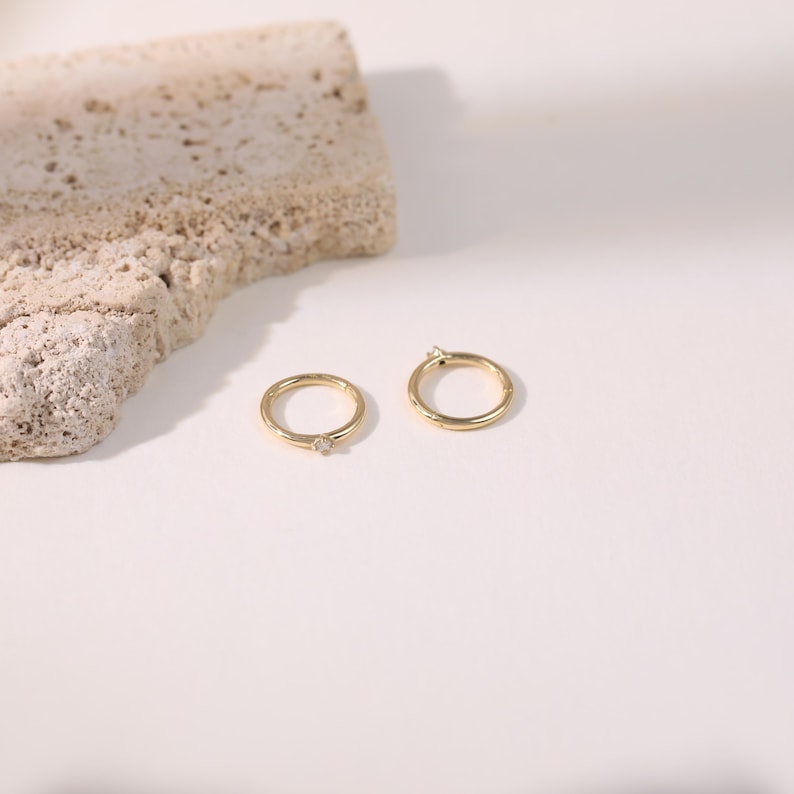 May include: Two gold hoop earrings with small, clear gemstones. One earring has a gemstone at the clasp, and the other has a gemstone in the center. The earrings are on a white surface with a textured, beige stone in the background.