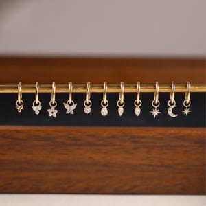 May include: A collection of gold hoop earrings with various charms, including a flower, butterfly, and teardrop shapes. Each earring features small, sparkling gemstones. The earrings are displayed on a gold bar against a dark wooden background.