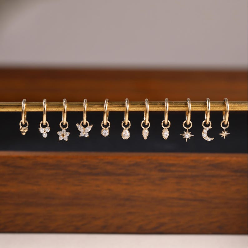May include: A collection of gold hoop earrings with various charms, including a flower, butterfly, and teardrop shapes. Each earring features small, sparkling gemstones. The earrings are displayed on a gold bar against a dark wooden background.